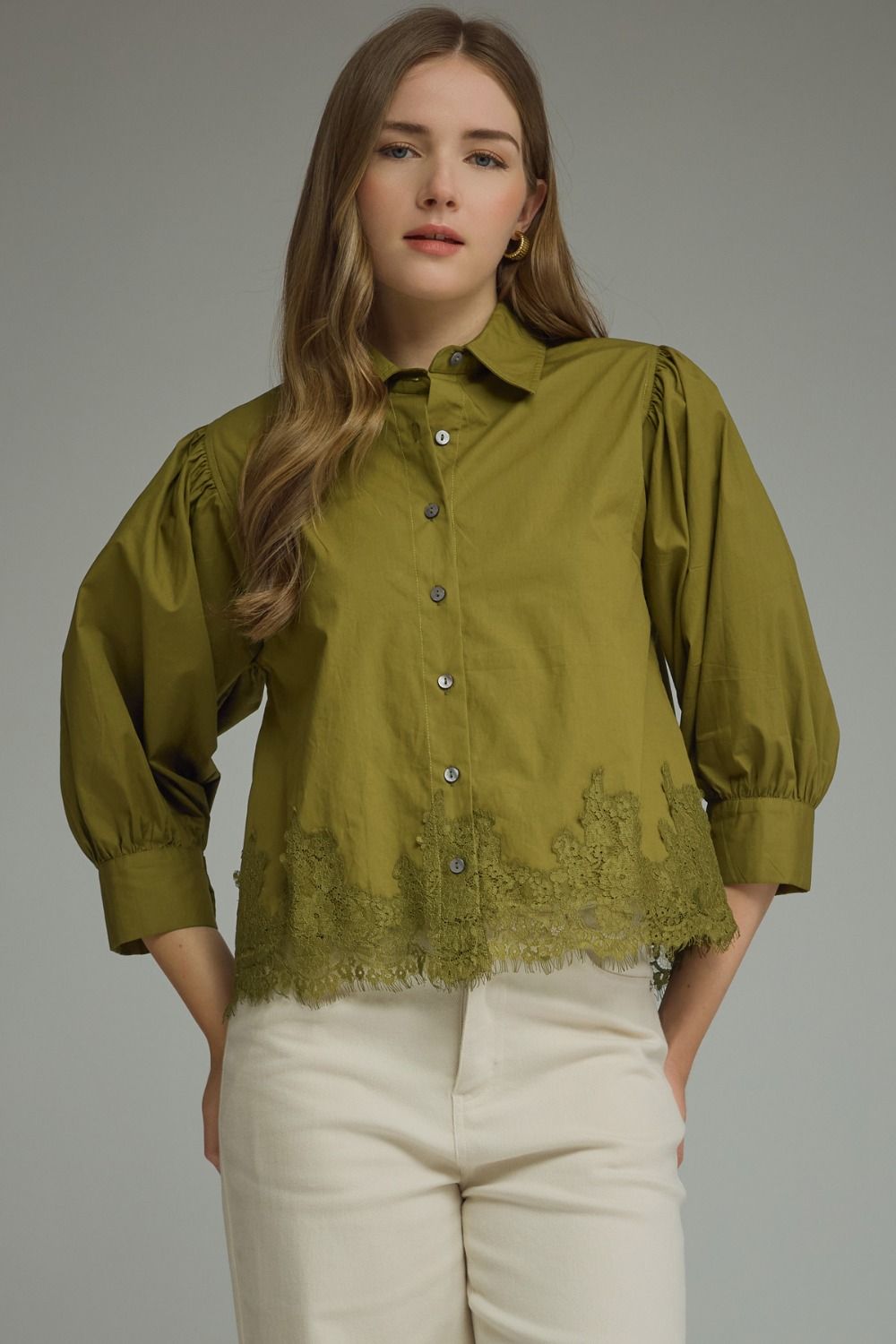 A woman wearing an olive green button-up blouse with puff sleeves and a lace hem, paired with cream-colored pants, against a solid gray studio background.