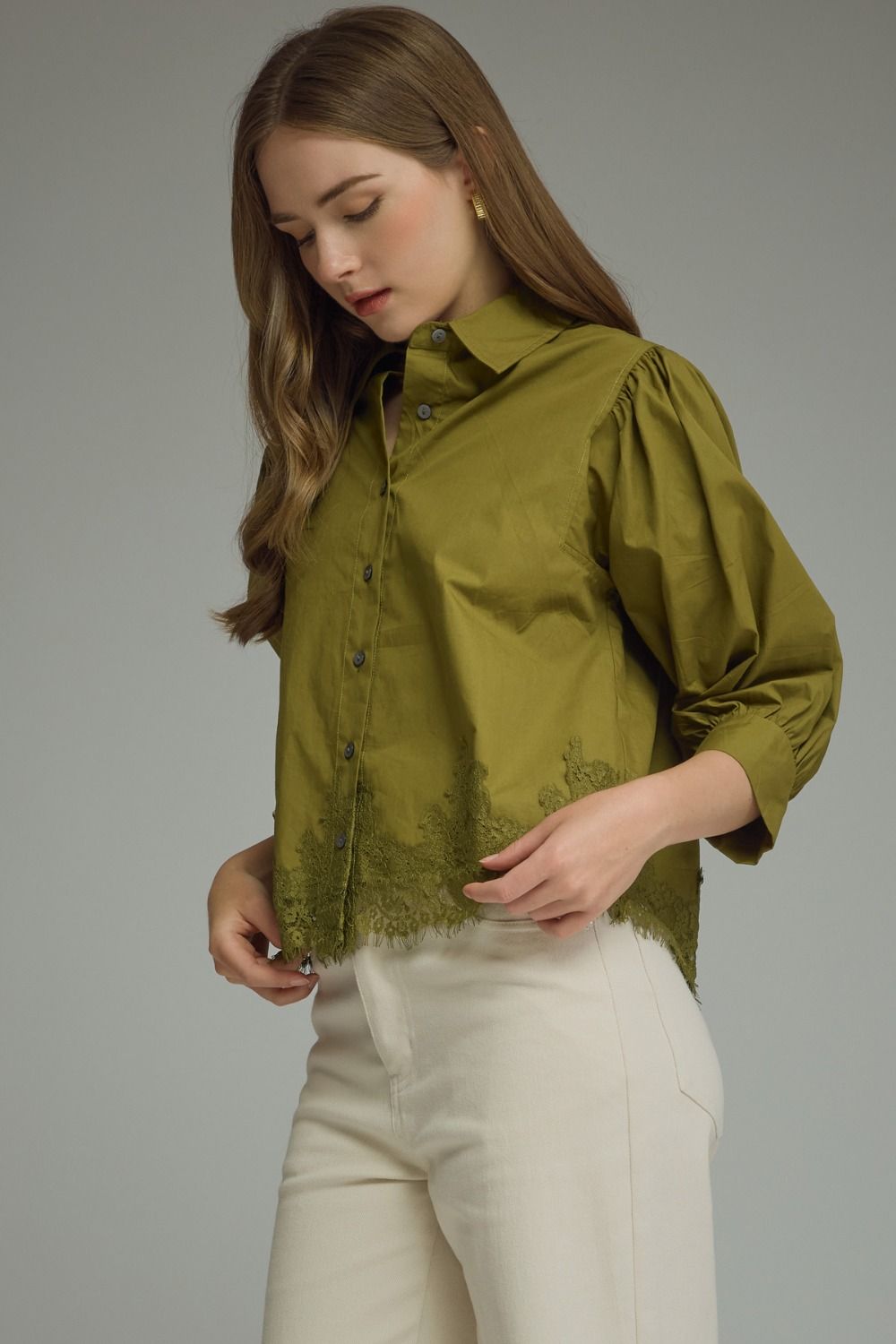 A woman wearing an olive green long-sleeve blouse with lace trim and cream-colored pants, photographed against a neutral gray studio background.