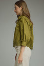 A woman shown in side profile wearing a cropped olive green blouse with voluminous puff sleeves and lace trim at the cuffs and hem, paired with light beige pants, against a plain gray studio background.