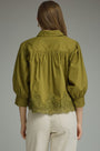 A woman viewed from the back wearing an olive green blouse with puffed three-quarter sleeves, gathered detailing at the shoulders, and a scalloped lace hem, paired with light beige trousers against a neutral gray background.