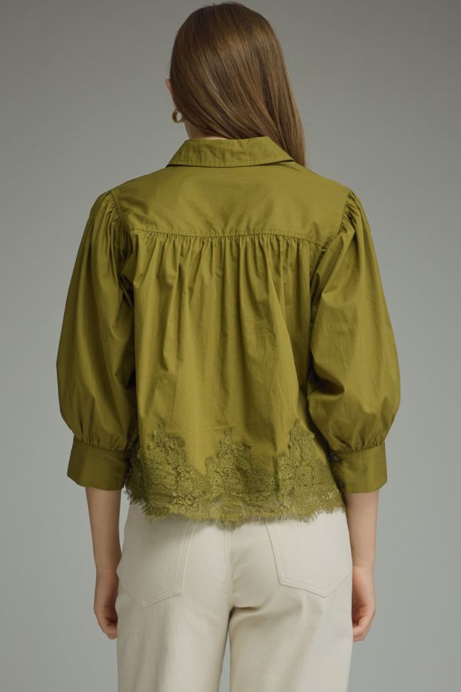 A woman viewed from the back wearing an olive green blouse with puffed three-quarter sleeves, gathered detailing at the shoulders, and a scalloped lace hem, paired with light beige trousers against a neutral gray background.