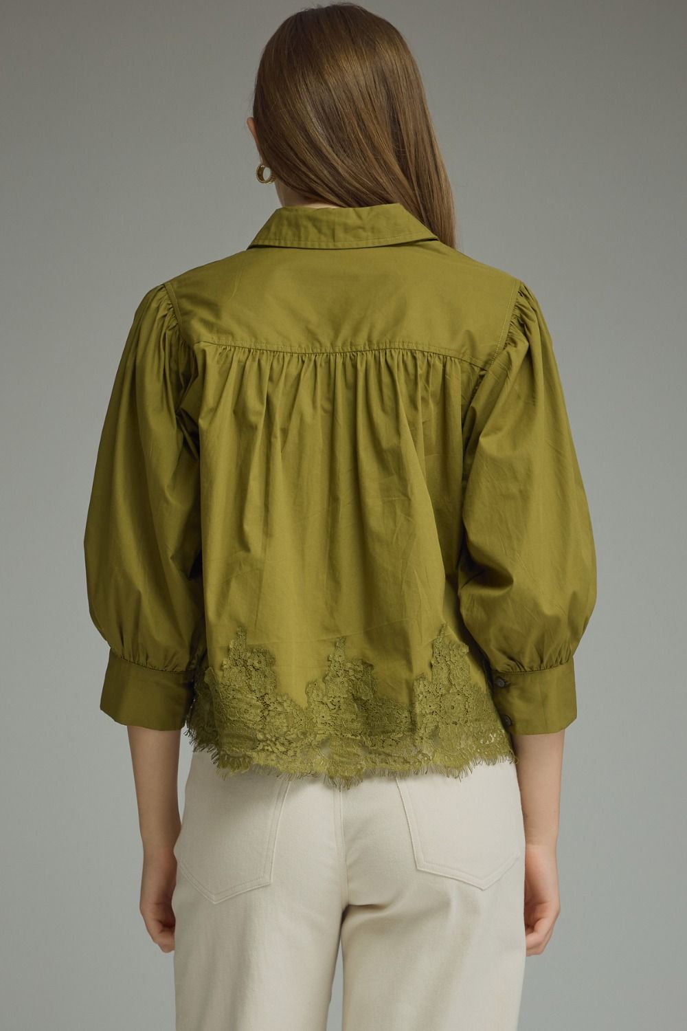 A woman viewed from the back wearing an olive green blouse with puffed three-quarter sleeves, gathered detailing at the shoulders, and a scalloped lace hem, paired with light beige trousers against a neutral gray background.