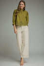 A woman modeling a sage green button-up blouse with lace trim at the hem and three-quarter sleeves, paired with off-white wide-leg trousers and beige heeled sandals, against a solid gray studio background.