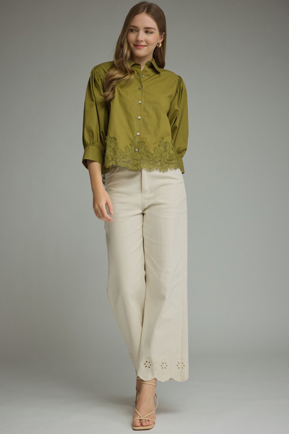 A woman modeling a sage green button-up blouse with lace trim at the hem and three-quarter sleeves, paired with off-white wide-leg trousers and beige heeled sandals, against a solid gray studio background.
