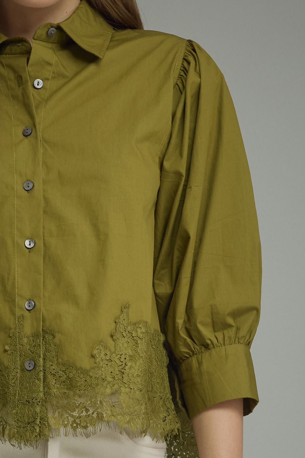An olive green button-up blouse with gathered puff sleeves, a pointed collar, and a matching lace hem, shown on a model against a neutral gray background.