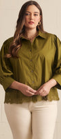 A woman modeling a short-sleeve olive green blouse with a button-down front, puffed shoulders, and a lace hem, paired with cream pants against a light beige brick wall.