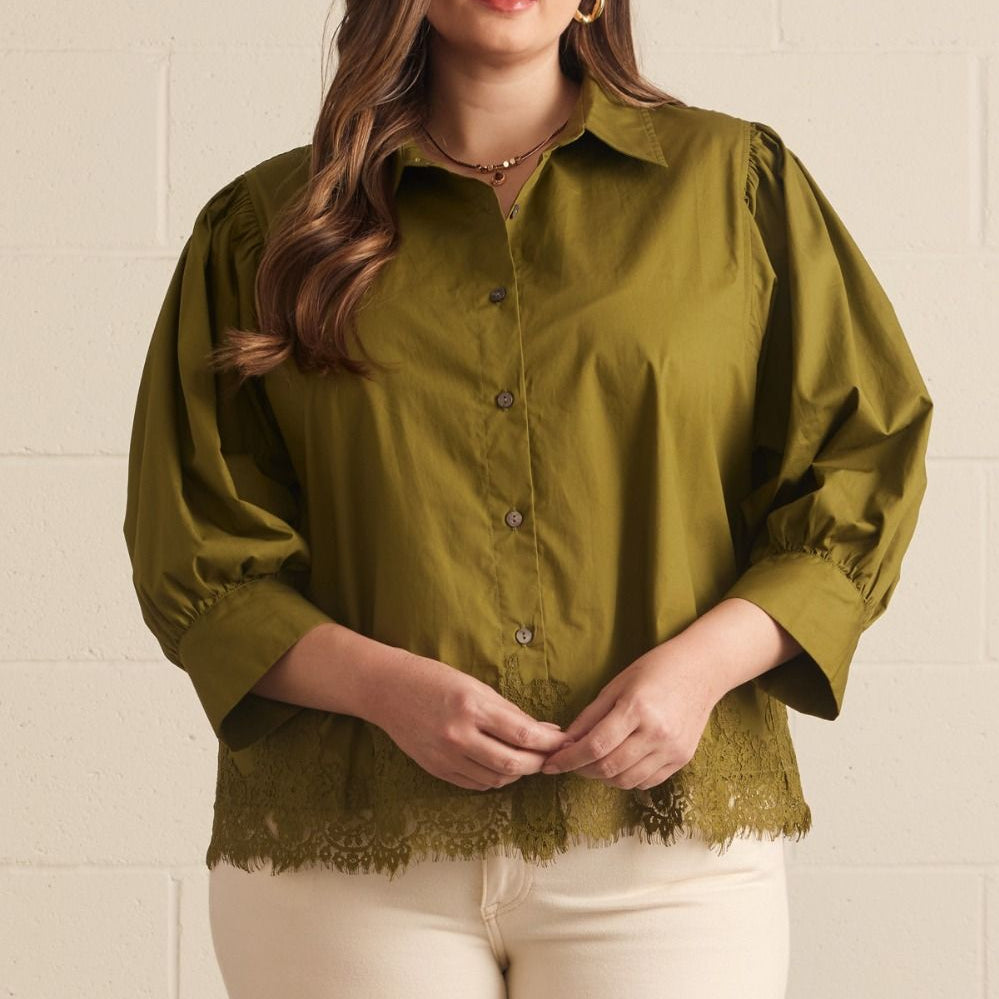 A woman modeling a short-sleeve olive green blouse with a button-down front, puffed shoulders, and a lace hem, paired with cream pants against a light beige brick wall.