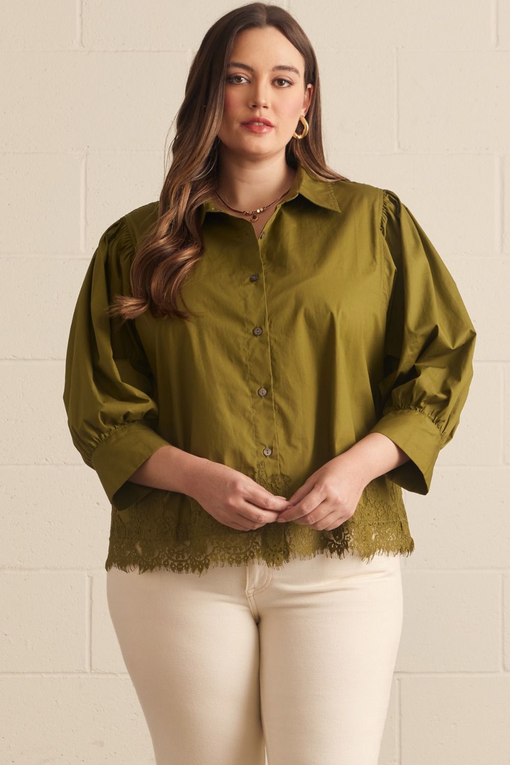 A woman modeling a short-sleeve olive green blouse with a button-down front, puffed shoulders, and a lace hem, paired with cream pants against a light beige brick wall.