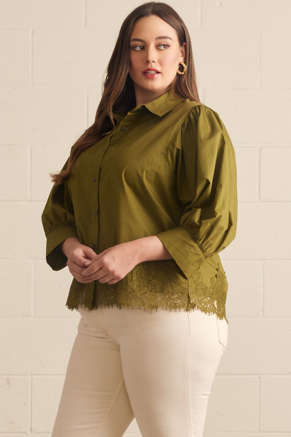 A woman modeling a sage green button-up blouse with puffed three-quarter sleeves and a lace hem, paired with cream pants against a light beige brick wall.