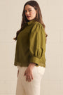 A woman wearing an olive green blouse with puffed three-quarter sleeves and lace trim at the cuffs and hem, paired with cream pants, standing against a white painted brick wall.