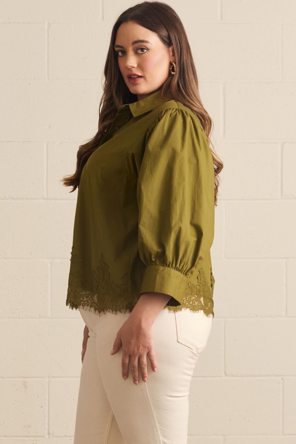 A woman wearing an olive green blouse with puffed three-quarter sleeves and lace trim at the cuffs and hem, paired with cream pants, standing against a white painted brick wall.