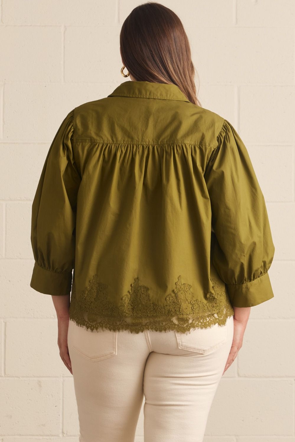 A woman viewed from the back wearing an olive green blouse with gathered puff sleeves and lace trim at the hem, paired with cream pants against a white brick wall.