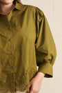 A close-up of a woman wearing an olive green button-up blouse with gathered puff sleeves and lace trim at the hem, photographed against a light neutral wall.