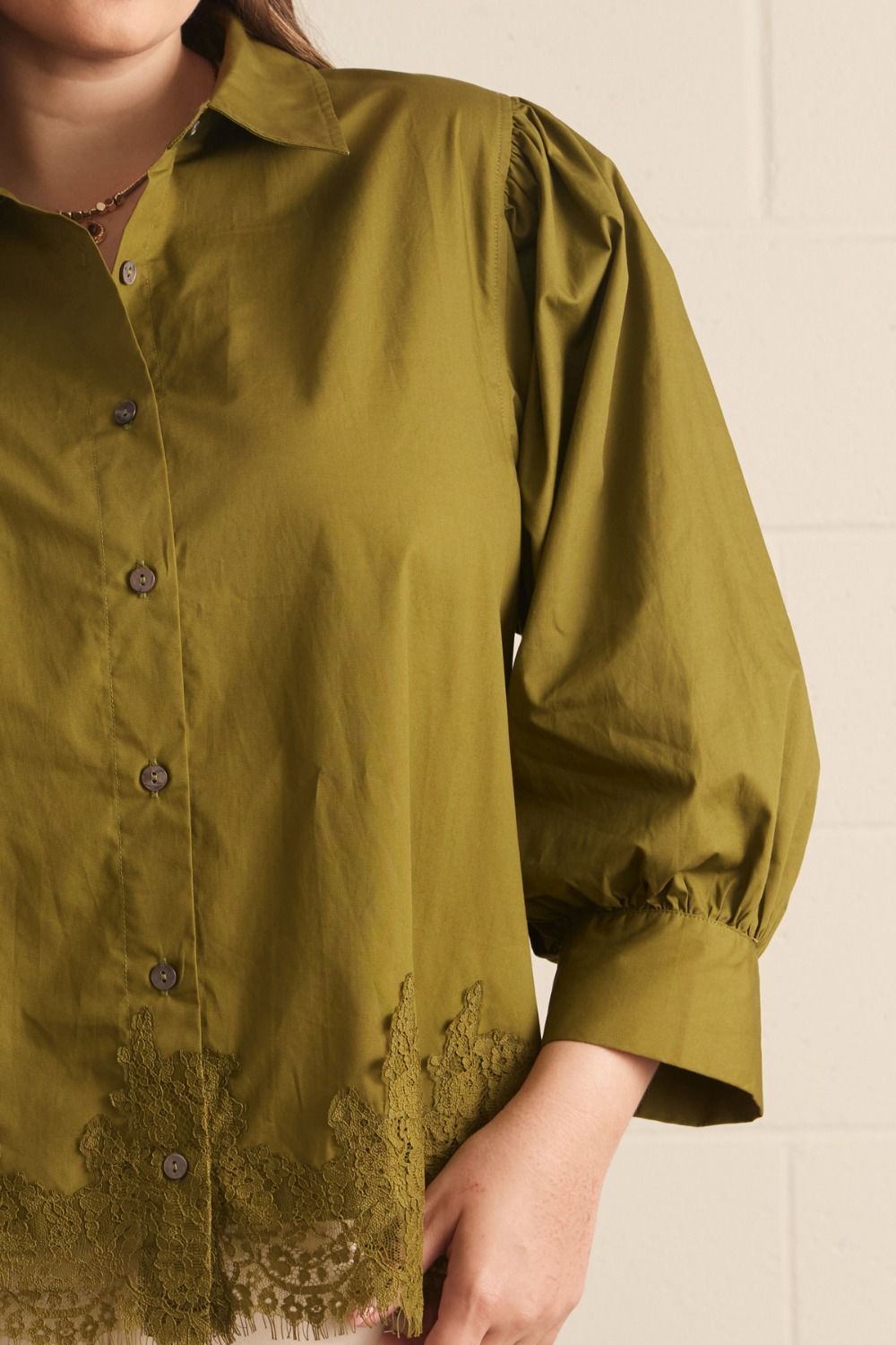 A close-up of a woman wearing an olive green button-up blouse with gathered puff sleeves and lace trim at the hem, photographed against a light neutral wall.