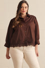 A woman modeling a dark brown button-up blouse with lace trim at the hem and three-quarter sleeves, paired with cream pants, standing against a light beige brick wall.