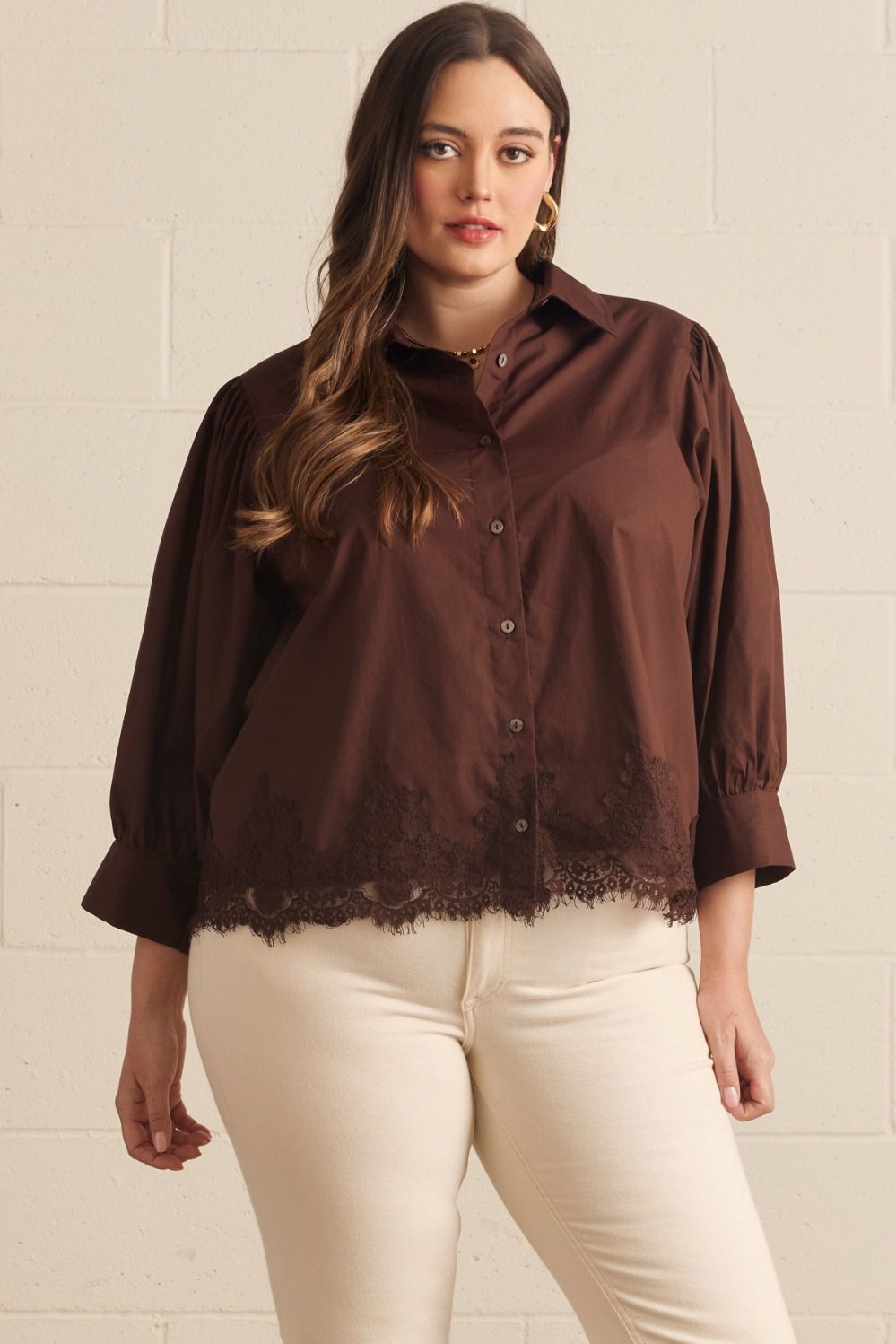 A woman modeling a dark brown button-up blouse with lace trim at the hem and three-quarter sleeves, paired with cream pants, standing against a light beige brick wall.