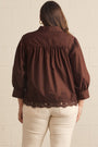 A woman viewed from the back wearing a dark brown blouse with gathered puff sleeves and lace trim at the hem, paired with cream pants, against a light brick wall.
