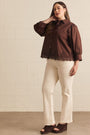 A woman modeling a brown long-sleeve button-up blouse with lace trim at the hem, paired with cream-colored pants and dark brown wedge sandals, standing against a light beige brick wall.