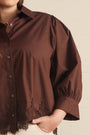 A close-up of a person wearing a brown button-front blouse with gathered puff sleeves and lace trim along the hem and cuffs, paired with light-colored bottoms against a plain light wall.