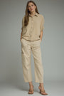 A woman modeling a beige short-sleeve button-up shirt and matching beige cropped cargo pants against a plain gray studio background.
