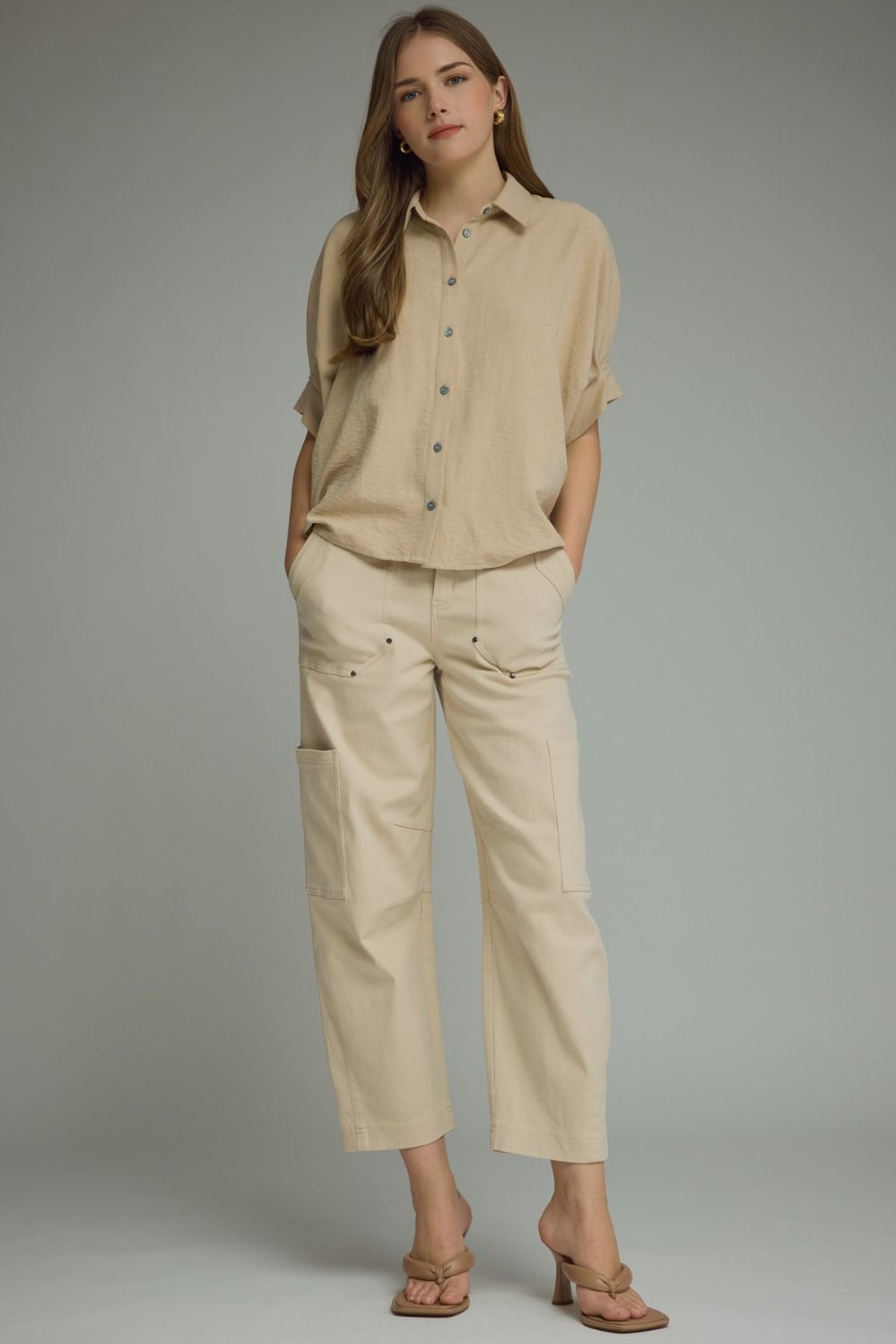A woman modeling a beige short-sleeve button-up shirt and matching beige cropped cargo pants against a plain gray studio background.