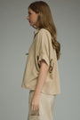 A woman in profile wearing a beige short-sleeve button-up shirt and matching beige trousers, photographed against a plain light gray studio background.
