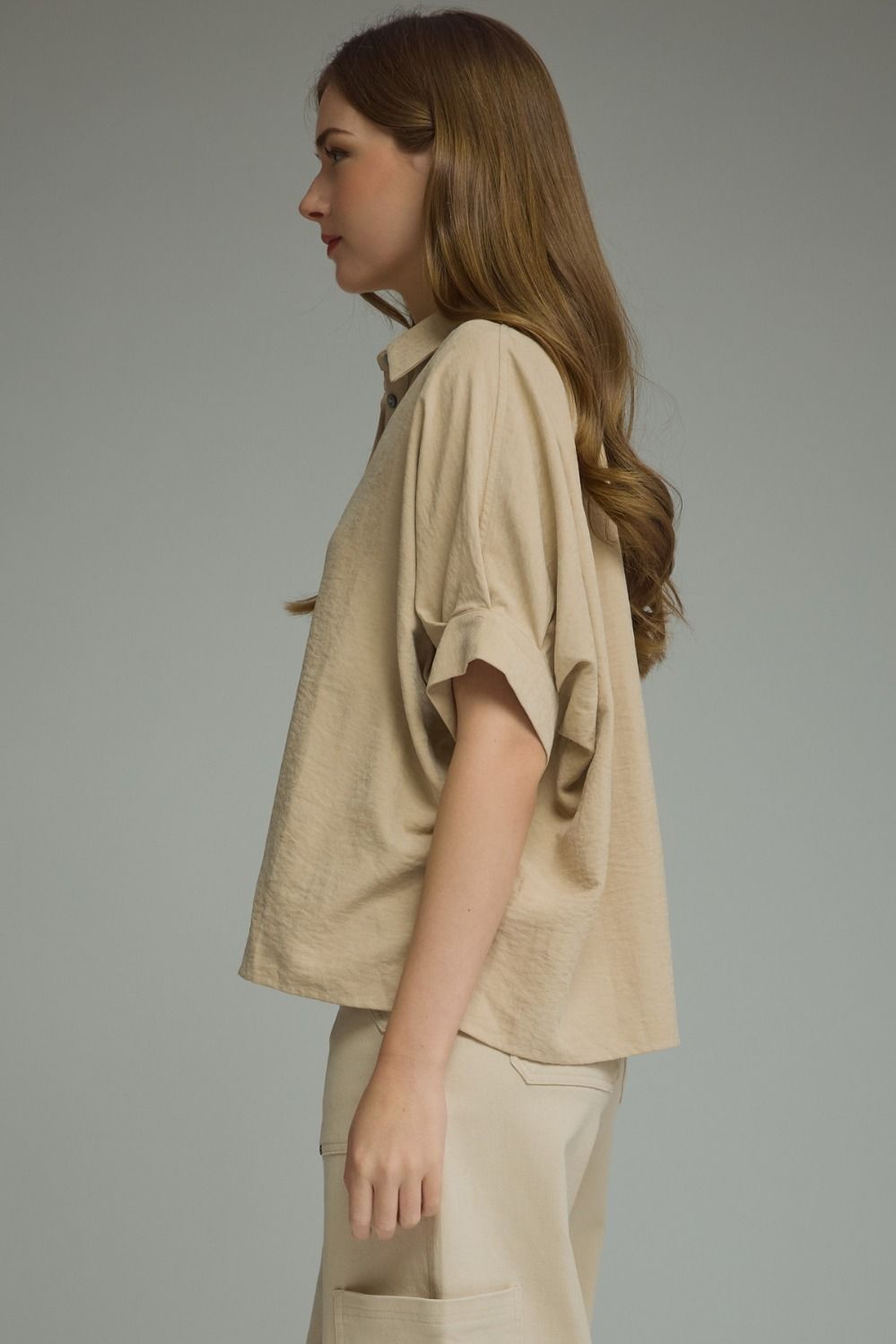 A woman in profile wearing a beige short-sleeve button-up shirt and matching beige trousers, photographed against a plain light gray studio background.