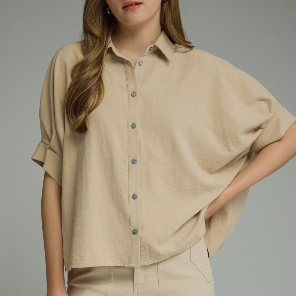 A woman modeling a beige short-sleeve button-up shirt with a relaxed, oversized fit and matching beige pants against a solid gray studio background.
