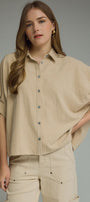 A woman modeling a beige short-sleeve button-up shirt with a relaxed, oversized fit and matching beige pants against a solid gray studio background.