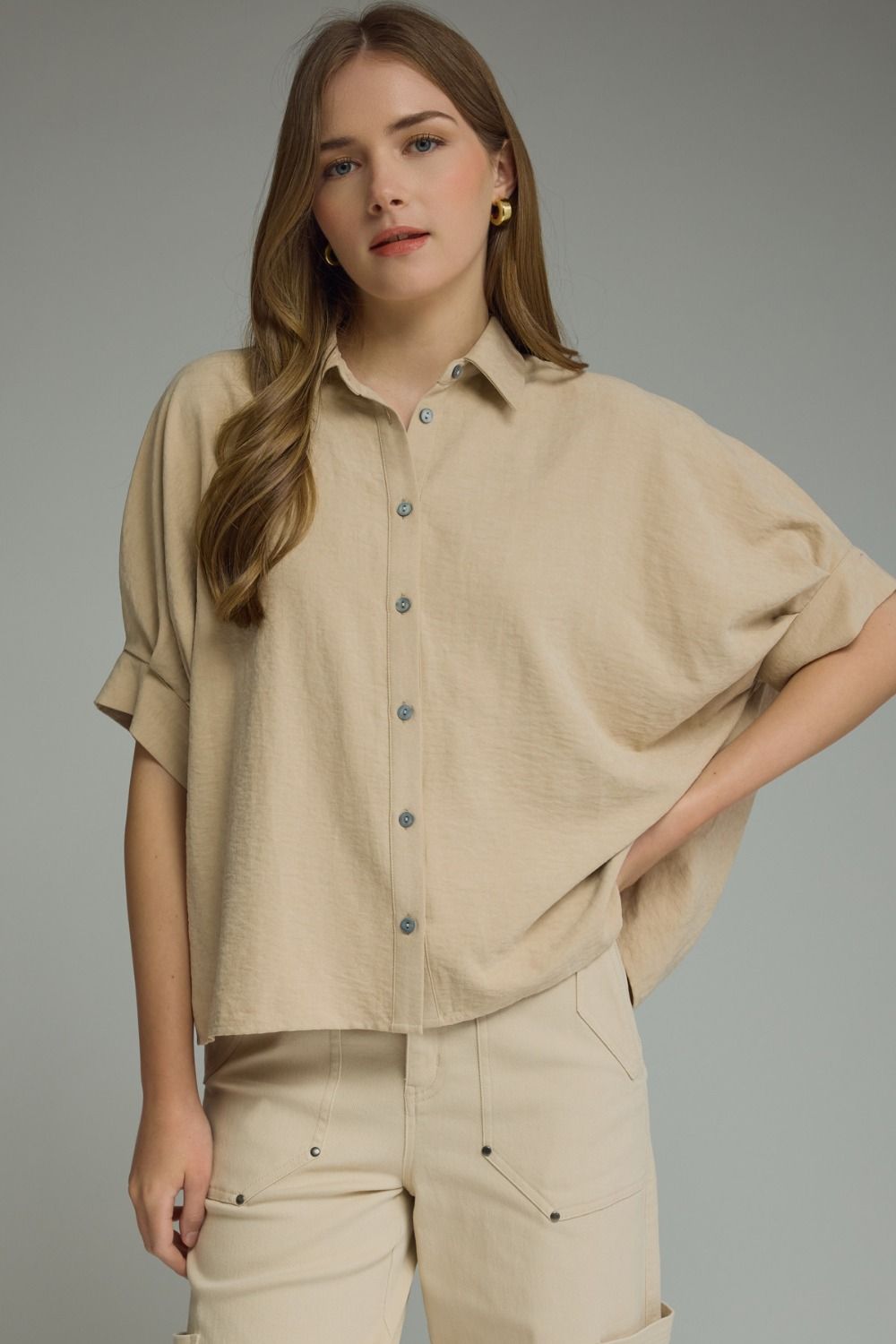 A woman modeling a beige short-sleeve button-up shirt with a relaxed, oversized fit and matching beige pants against a solid gray studio background.