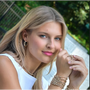 Woman with blonde hair and pink lipstick wearing multiple bracelets and rings outdoors.