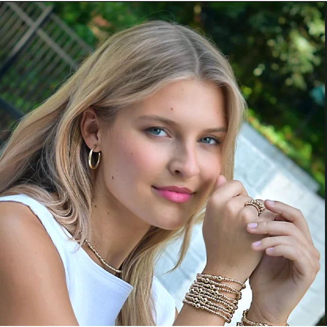 Woman with blonde hair and pink lipstick wearing multiple bracelets and rings outdoors.