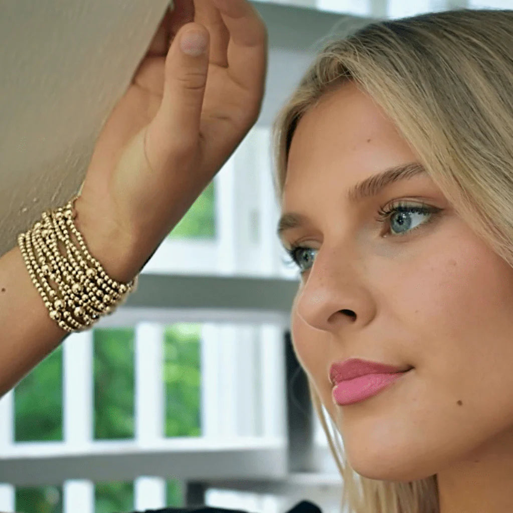Woman with pink lipstick and blonde hair, wearing a gold bracelet, with a blurred background.