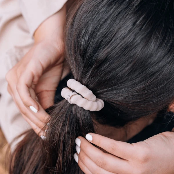 A close-up lifestyle shot of a person with dark brown hair wearing a ribbed, light beige scrunchie. Their hands are gently adjusting the hair tie, and the background is softly blurred.