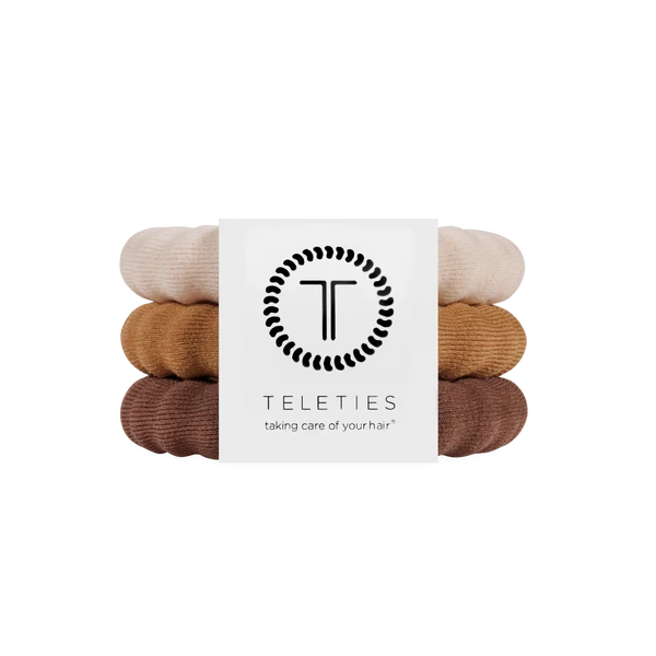 Three Teleties hair tie bands stacked vertically with a white paper label in the center. The bands are in three different neutral shades: a light cream, a warm camel tan, and a medium brown.