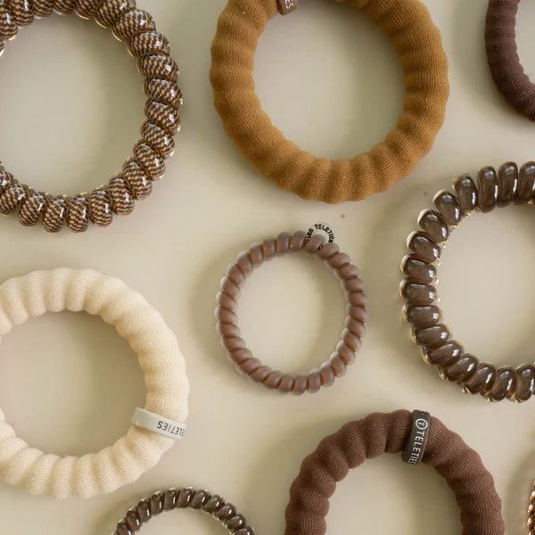 An assortment of fabric and elastic hair ties in various colors and textures, including brown, beige, and cream, arranged on a light cream background.