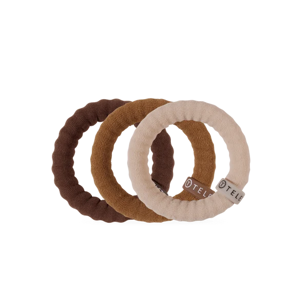 Three textured fabric baby teething rings in dark brown, medium brown, and cream, overlapping on a solid white background. The cream ring features a small silver metal brand tag with the word 'TELL'.