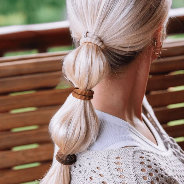 A close-up back view of a woman with blonde hair styled in a low ponytail using three different brown hair ties, seated on a wooden bench.