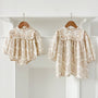 Two floral baby outfits, a dress and a romper, hanging on wooden hangers against a white wall.