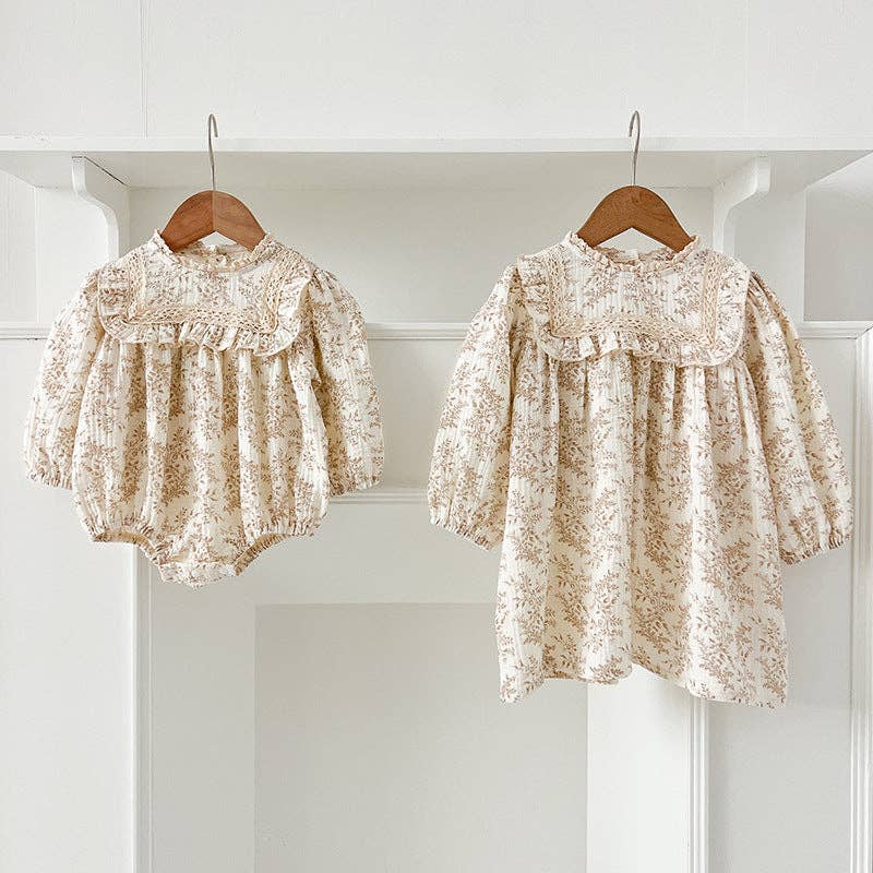 Two floral baby outfits, a dress and a romper, hanging on wooden hangers against a white wall.