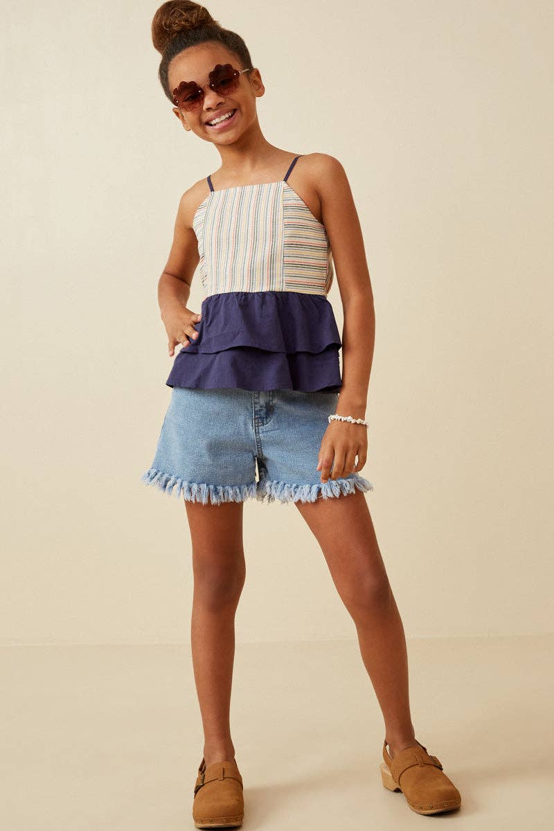 Young girl wearing a layered top and denim shorts with sunglasses on a beige background