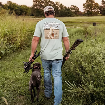 Man in a field with a dog, holding hunting rifles, wearing a 'Fiststone' t-shirt.
