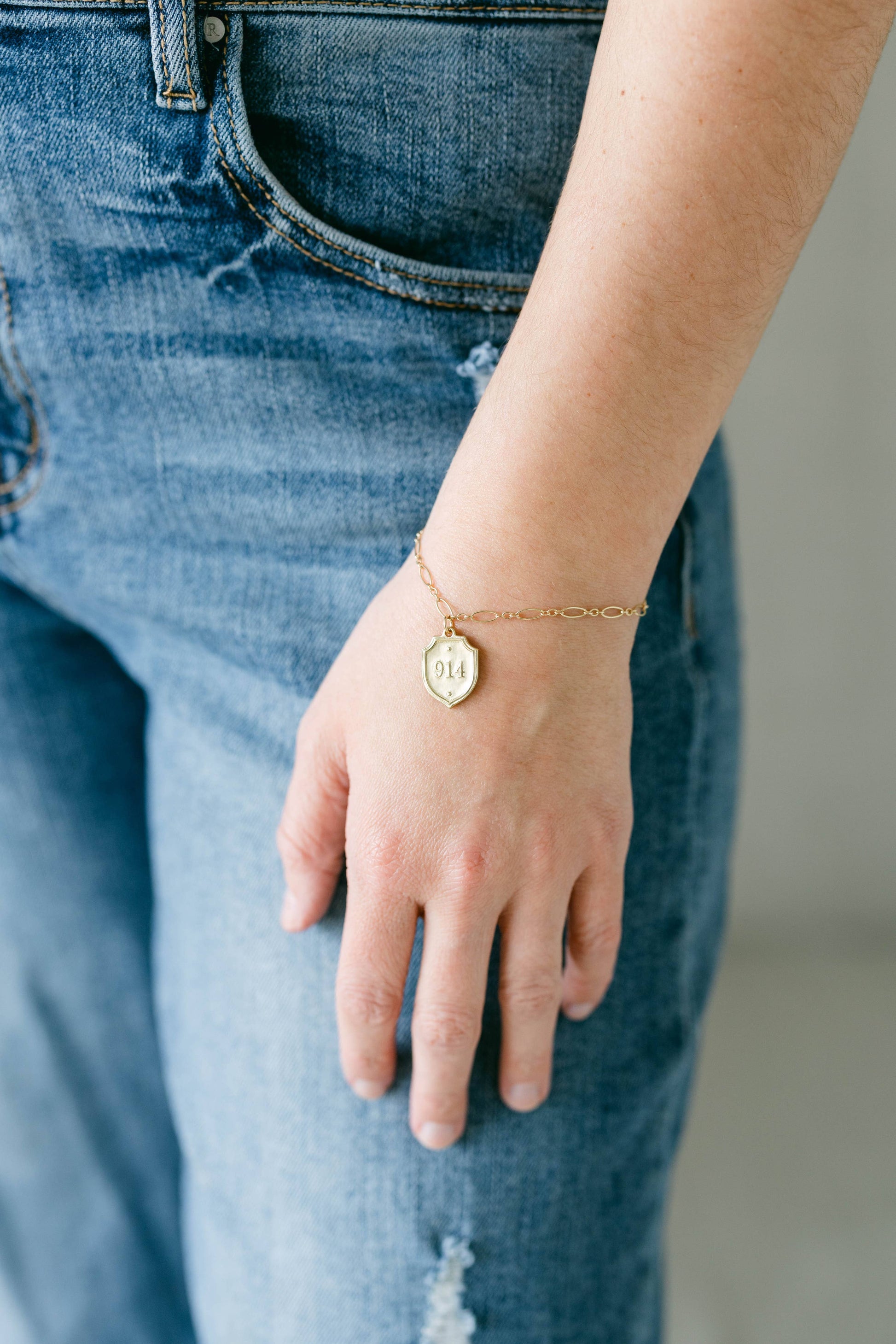 A close-up of a person's hand resting on their denim jeans, showcasing a delicate gold chain bracelet with a small oval charm 