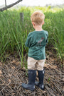 Child wearing a green shirt and navy rain boots standing in a natural setting with tall grass.