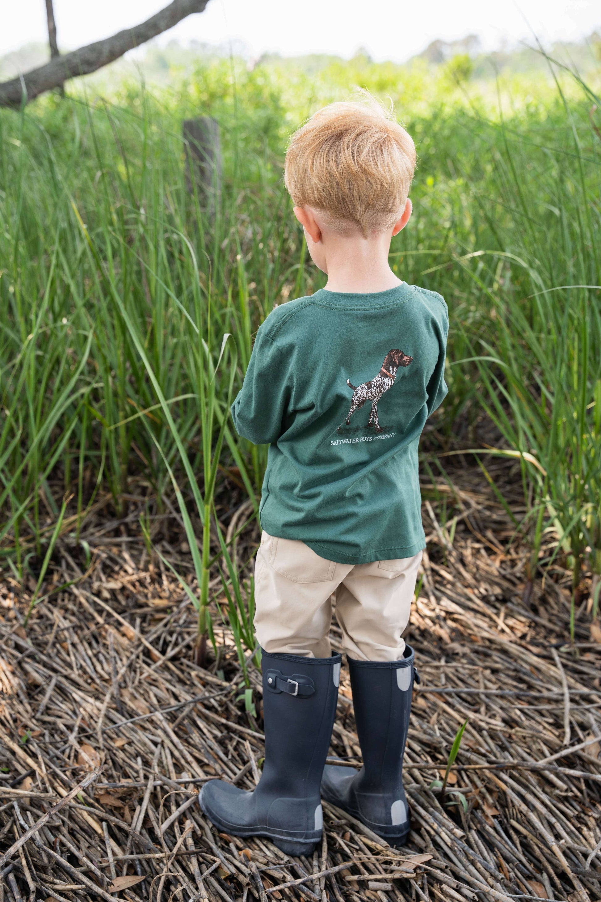 Child wearing a green shirt and navy rain boots standing in a natural setting with tall grass.