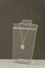 A gold chain necklace with a round engraved pendant displayed on a clear acrylic stand against a textured dark gray background.