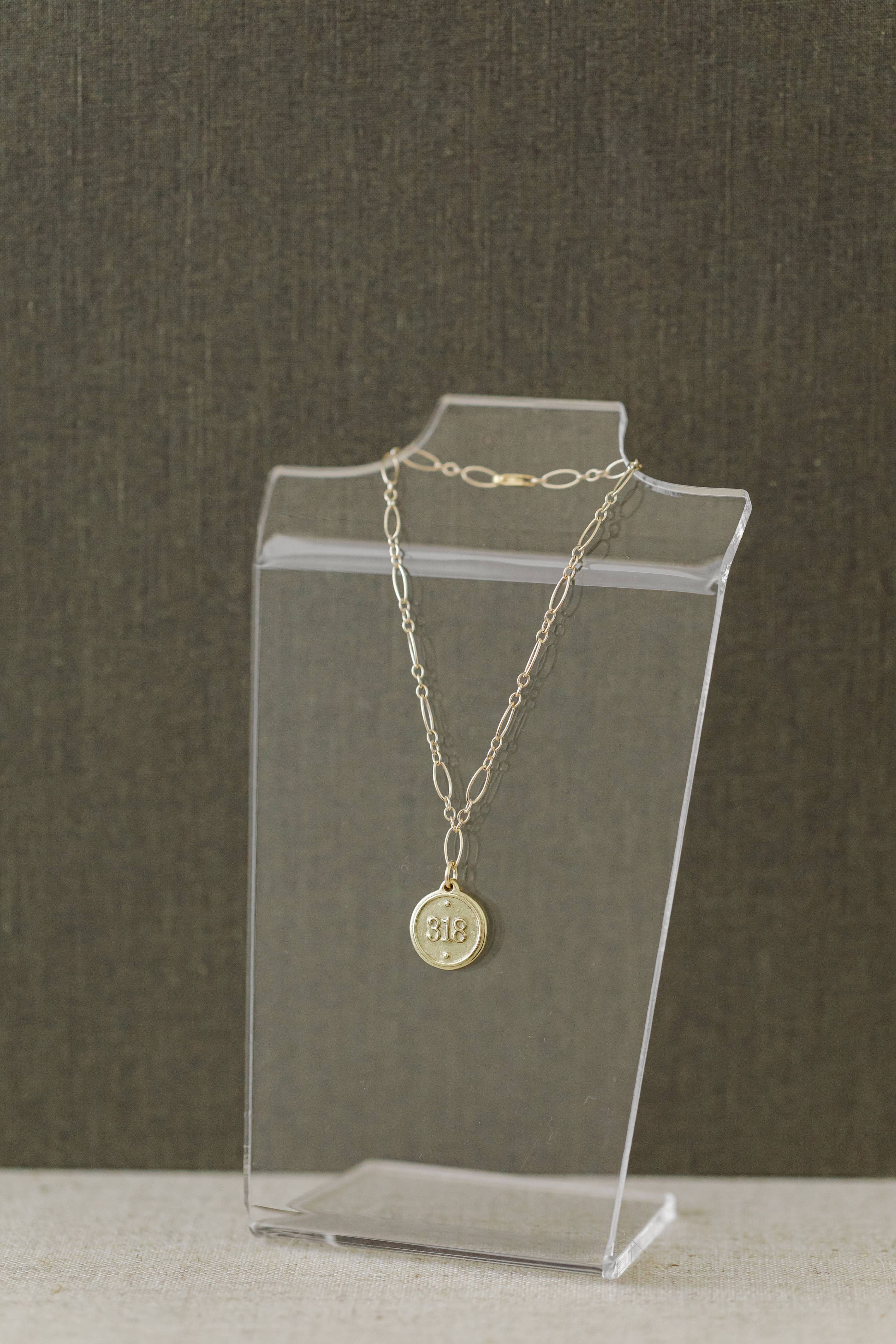 A gold chain necklace with a round engraved pendant displayed on a clear acrylic stand against a textured dark gray background.