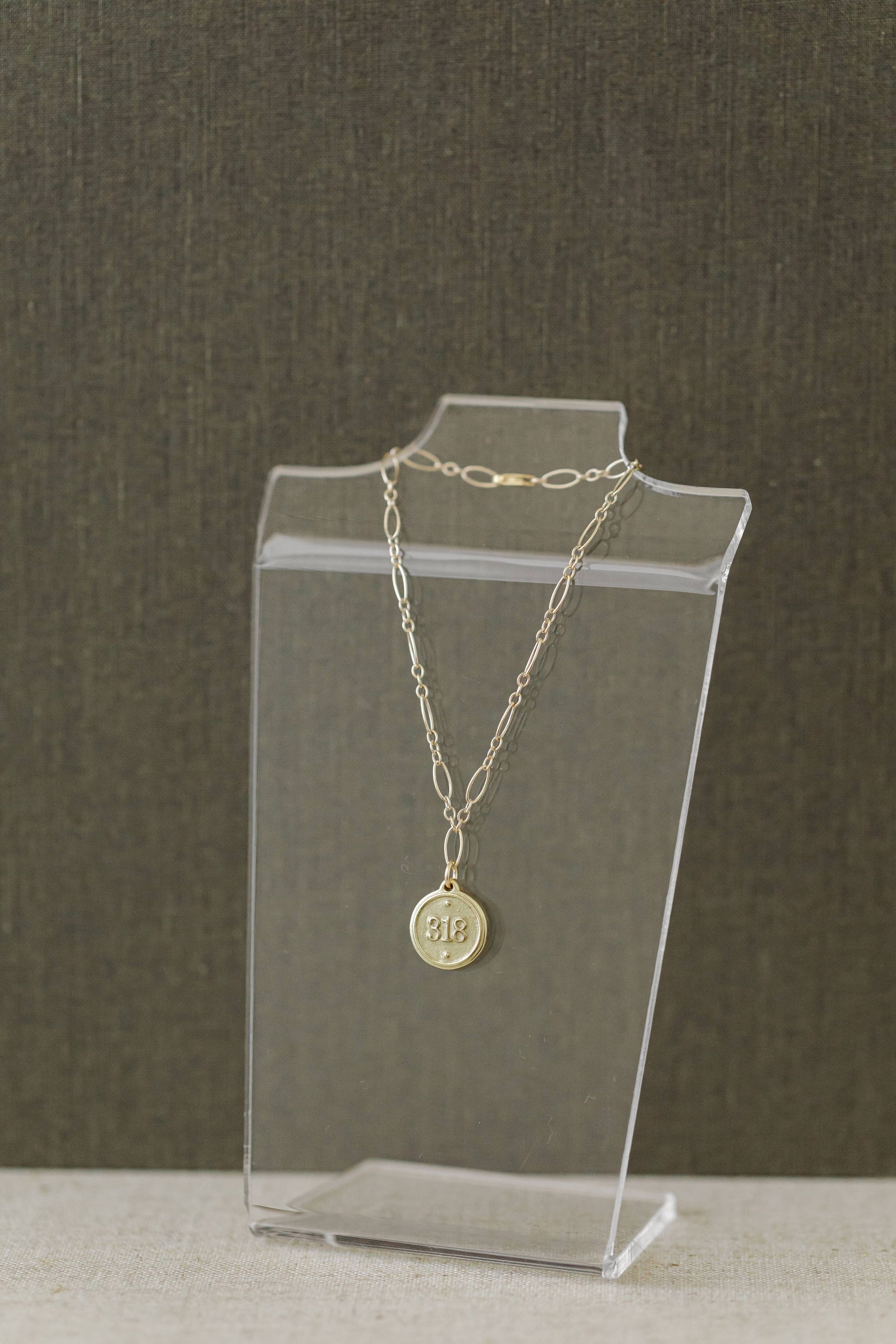 A gold chain necklace with a round engraved pendant displayed on a clear acrylic stand against a textured dark gray background.