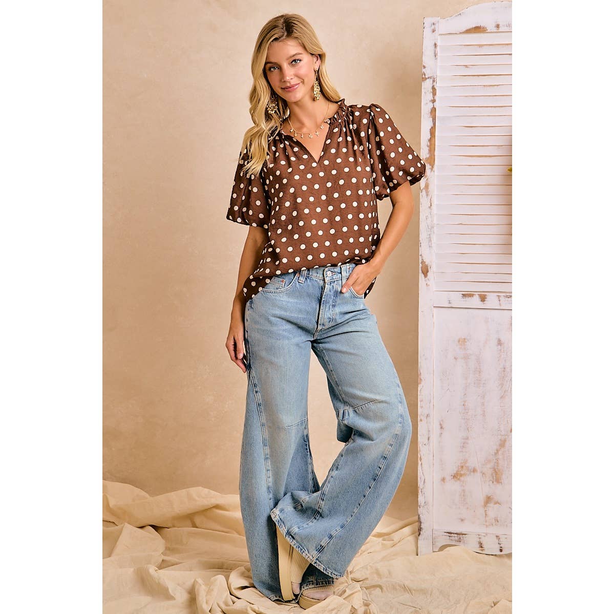 Woman wearing a brown polka dot blouse and blue jeans against a beige wall.