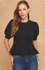 A woman modeling a black short-sleeve puff-sleeve top with a peplum hem, paired with blue jeans, against a textured tan background.